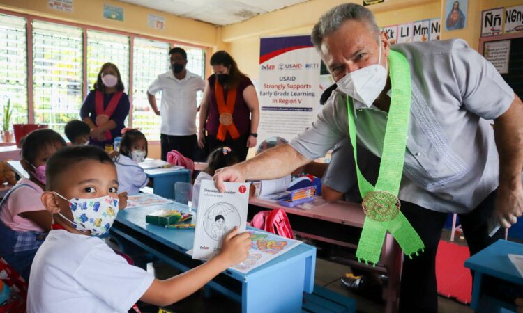 USAID Philippines Education Director Dr. LeBlanc hands a book to a grade 1 student of Salvacion Elementary School in Albay on the first day of classes in the Philippines.