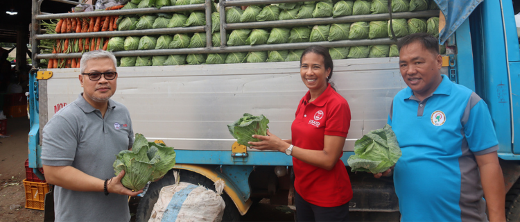 People holding cabbages in front of a truck carrying fresh produce.