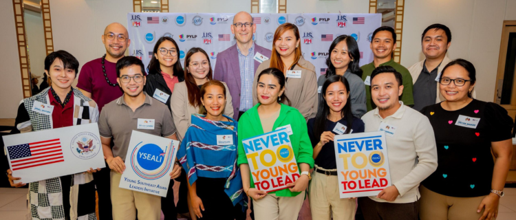 Deputy Chief of Mission Robert Ewing and U.S. government exchange program participants on stage while holding banners.